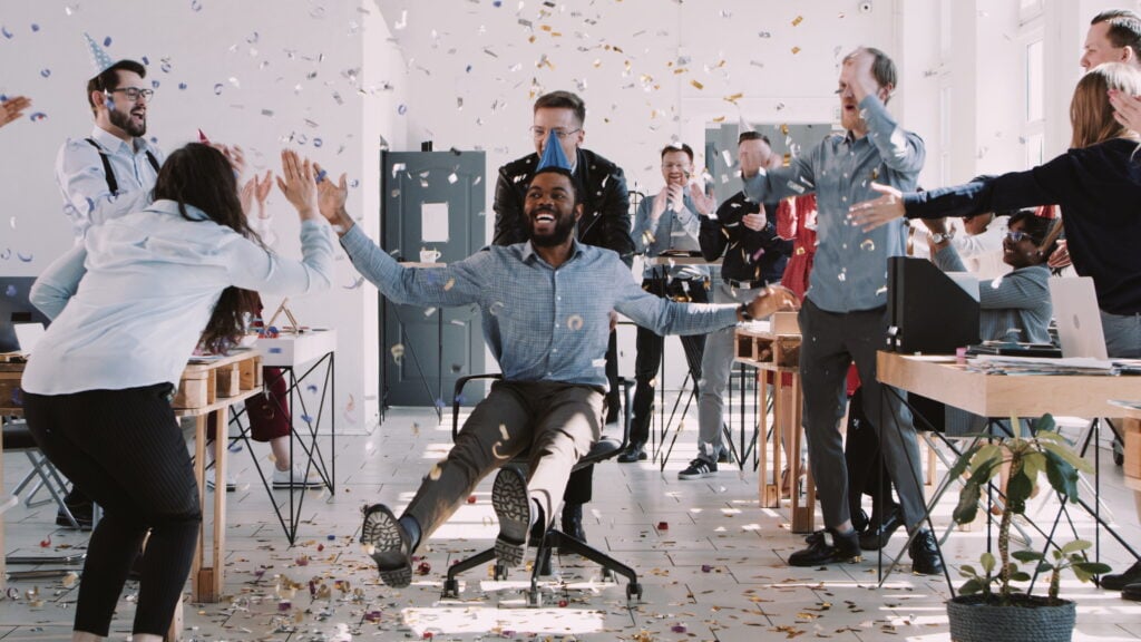 Employees wheeling person in office chair and giving birthday wishes to boss